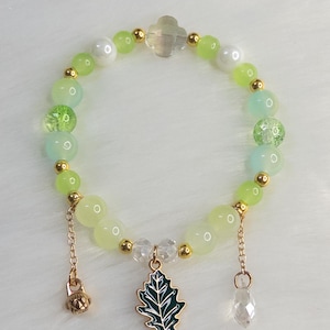 May include: A beaded bracelet featuring a mix of green, white, and gold beads. The bracelet has a gold charm with a green leaf design and a dangling clear crystal. The beads are various sizes and shades of green.