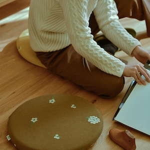 以下が含まれることがあります： 白い花の刺繍が施された丸い茶色のクッション。クッションは木の床にあります。黄色いクッションに座り、白いセーターを着ている人がいます。木製の靴型と製図板も床にあります。