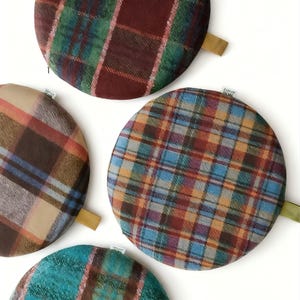 May include: A collection of round, plaid fabric items in various colors. The patterns include shades of red, green, blue, brown, and tan. Each item has a small, attached fabric loop. The items are arranged on a white background.