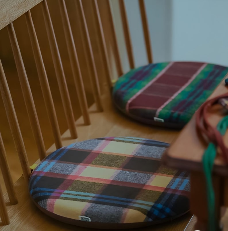 May include: Two round seat cushions with a plaid pattern in shades of brown, blue, pink, and yellow rest on a wooden chair. The cushions are placed on a light-colored wooden surface. The chair has a slatted back.