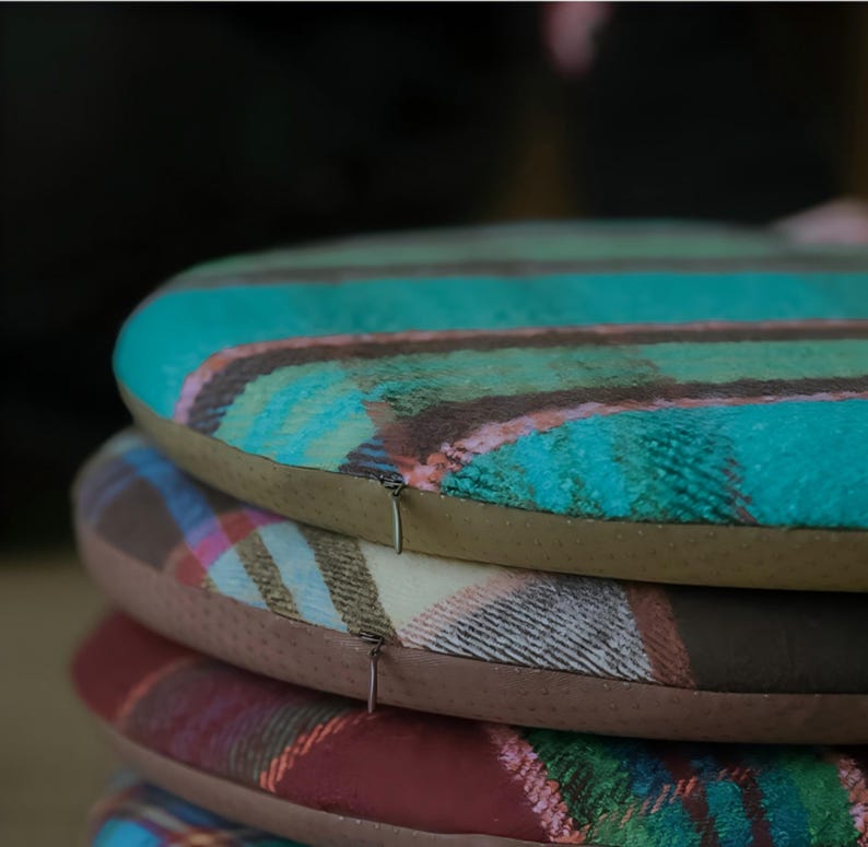 May include: A stack of four round seat cushions with plaid patterns. The top cushion is teal, brown, and pink. The other cushions are brown, cream, and red. Each cushion has a brown border and a zipper.
