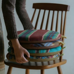 May include: A stack of four round chair cushions with colorful plaid patterns. The cushions are in shades of red, green, blue, and brown. Each cushion has a yellow fabric loop attached to the side. The cushions are placed on a wooden chair.