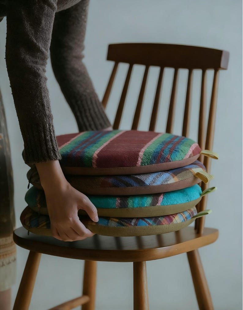 May include: A stack of four round chair cushions with colorful plaid patterns. The cushions are in shades of red, green, blue, and brown. Each cushion has a yellow fabric loop attached to the side. The cushions are placed on a wooden chair.