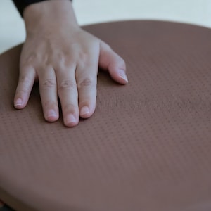 May include: A brown, circular exercise balance board with a textured surface. The board is designed for stability and core training. The image shows a hand resting on the board, highlighting its size and texture.