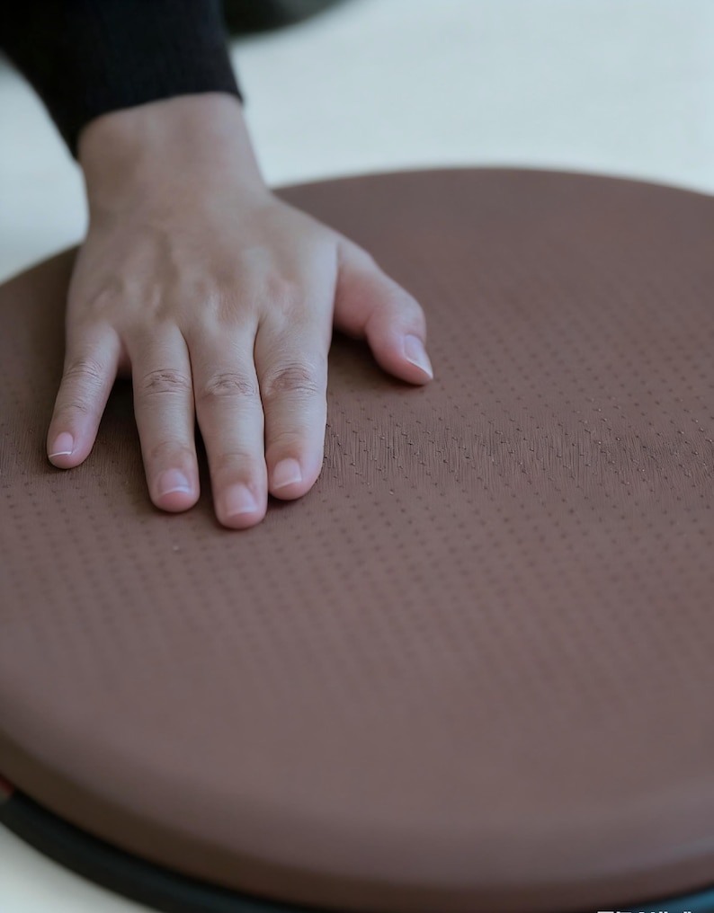 May include: A brown, circular exercise balance board with a textured surface. The board is designed for stability and core training. The image shows a hand resting on the board, highlighting its size and texture.