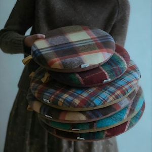 May include: A stack of round, plaid cushions in shades of brown, red, blue, and green. Each cushion has a contrasting brown border and a small tag. The cushions are held by a person wearing a brown sweater and dress.