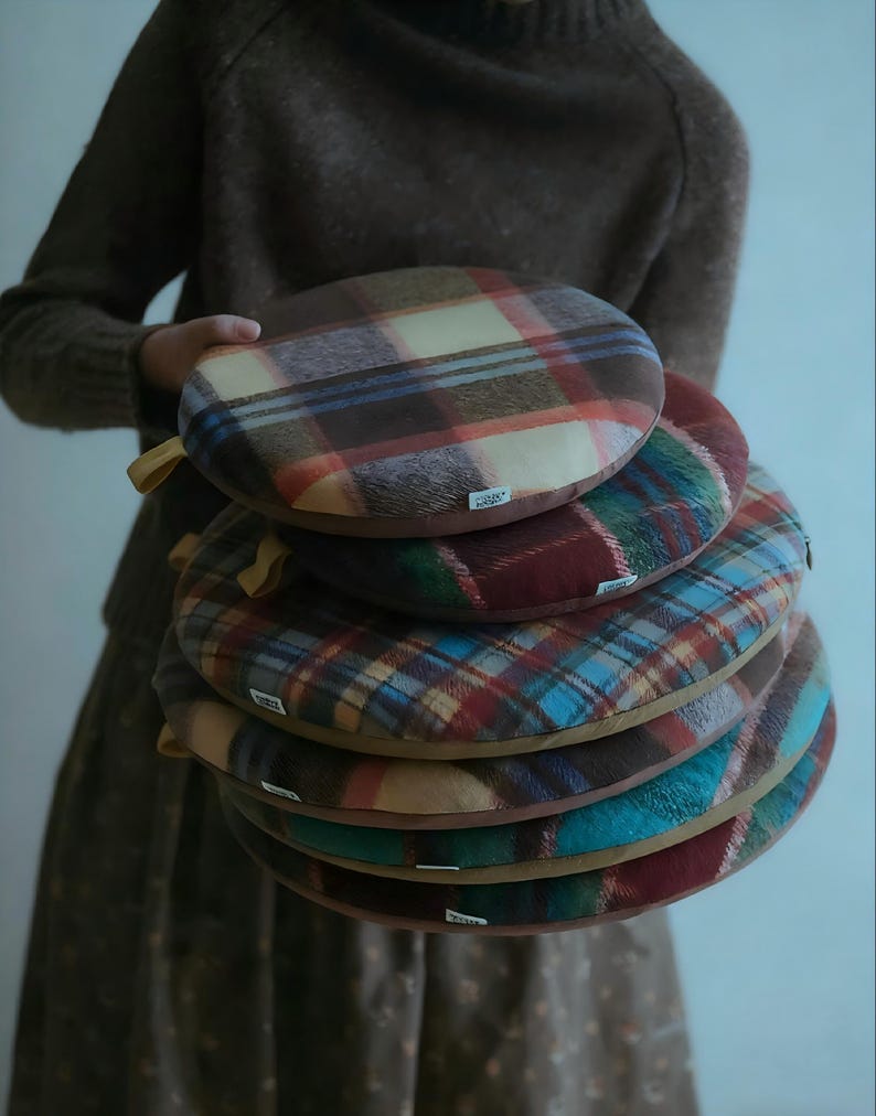 May include: A stack of round, plaid cushions in shades of brown, red, blue, and green. Each cushion has a contrasting brown border and a small tag. The cushions are held by a person wearing a brown sweater and dress.
