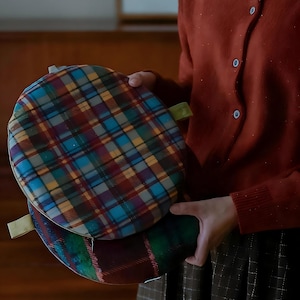 May include: Two round, plaid fabric items in shades of blue, red, yellow, and green. Each has a small loop attached. The items are held by a person wearing a rust-colored cardigan.