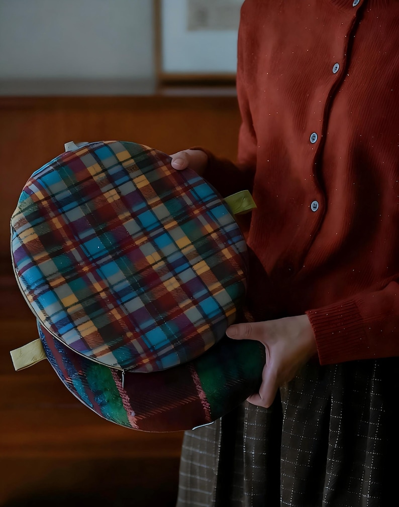 May include: Two round, plaid fabric items in shades of blue, red, yellow, and green. Each has a small loop attached. The items are held by a person wearing a rust-colored cardigan.
