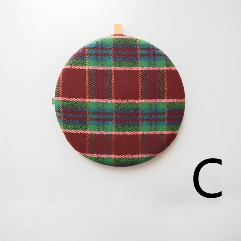 May include: A round, plaid trivet in shades of burgundy, green, and blue. The trivet has a wooden handle at the top. The trivet is a kitchen accessory.