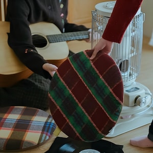 May include: Round cushion with a plaid pattern in burgundy, green, and white. The cushion is held by a person. Another cushion with a similar pattern is visible in the background. A guitar and a heater are also in the background.