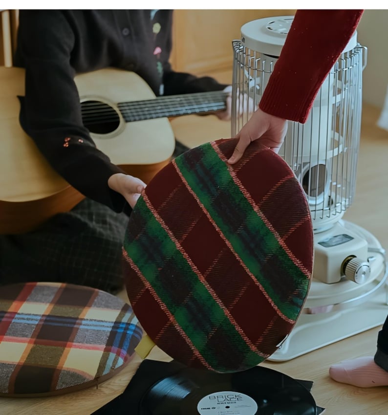May include: Round cushion with a plaid pattern in burgundy, green, and white. The cushion is held by a person. Another cushion with a similar pattern is visible in the background. A guitar and a heater are also in the background.