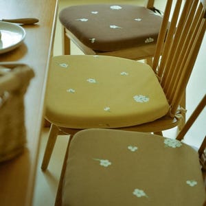 Op de afbeelding: Drie houten stoelen met bloempatroon zitkussens. De kussens zijn in bruin- en geeltinten, versierd met witte bloemenborduursels. De stoelen staan bij een houten tafel.