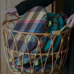 May include: A wicker basket filled with several plaid cushions in shades of brown, yellow, blue, and green. The cushions have a rounded shape and are stacked inside the basket. The basket has a handle.
