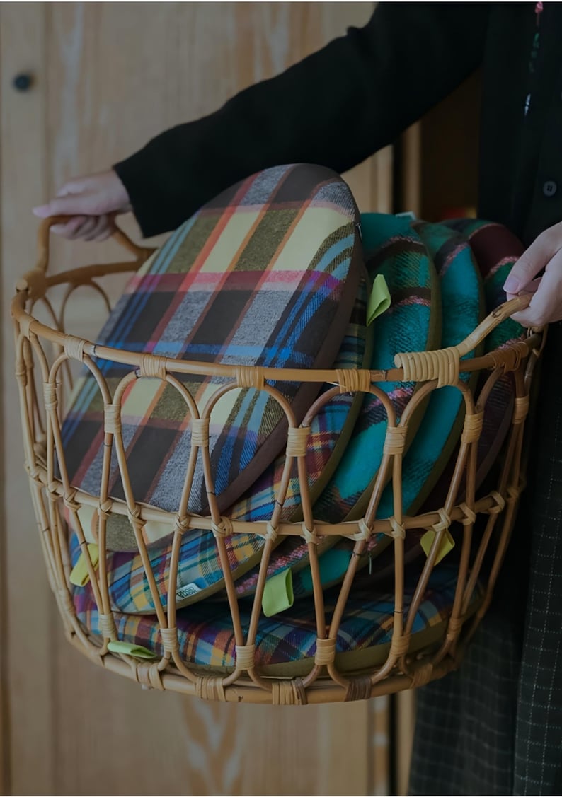 May include: A wicker basket filled with several plaid cushions in shades of brown, yellow, blue, and green. The cushions have a rounded shape and are stacked inside the basket. The basket has a handle.