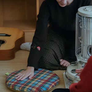 May include: A round, plaid cushion in red, blue, yellow, and green. The cushion is on a wooden floor. A person wearing a black sweater with embroidered forest and mushroom designs on the sleeve is touching the cushion. A guitar and a heater are in the background.