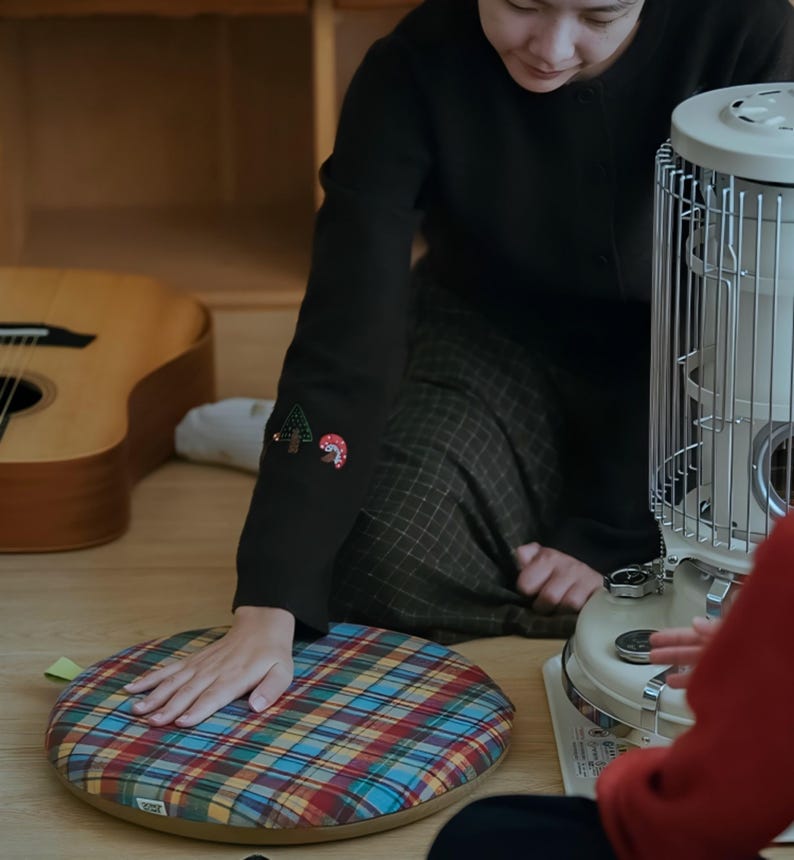 May include: A round, plaid cushion in red, blue, yellow, and green. The cushion is on a wooden floor. A person wearing a black sweater with embroidered forest and mushroom designs on the sleeve is touching the cushion. A guitar and a heater are in the background.