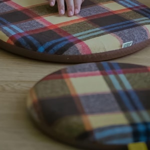 May include: Two round seat cushions with a plaid pattern in shades of brown, yellow, red, and blue. The cushions have a brown border and a yellow fabric loop. One cushion is in focus, while the other is slightly blurred.