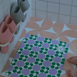 May include: A rectangular bath mat featuring a repeating pattern of green and lilac circles on a white backdrop. The mat rests on a tiled floor with pink and white tiles. Grey and pink slippers are on a rack.