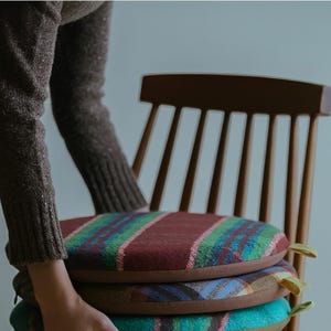 May include: A stack of three round seat cushions with colorful plaid patterns. The cushions are stacked on a wooden chair. The top cushion has a burgundy background with green and pink stripes. The second cushion has a brown background with a blue and green plaid pattern. The bottom cushion has a teal background with a plaid pattern.