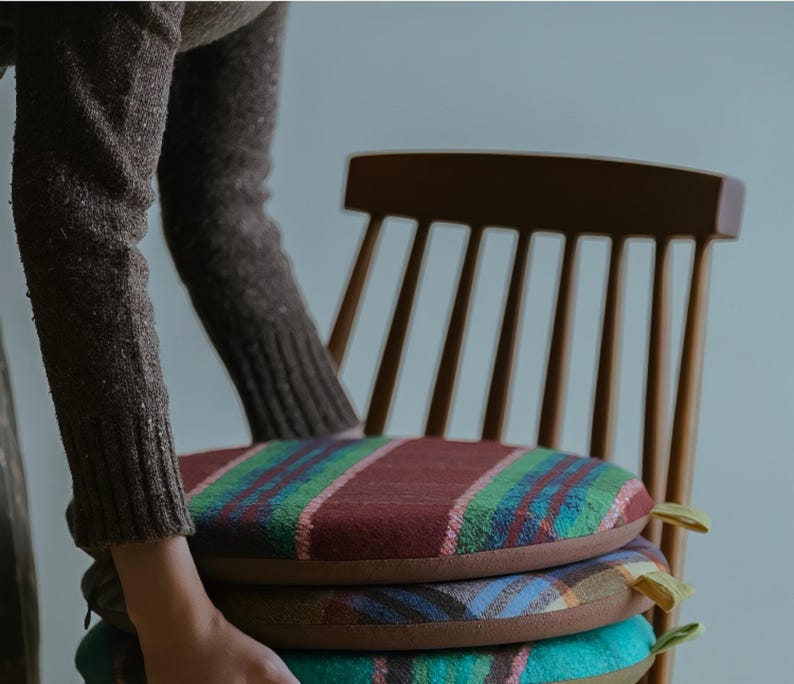 May include: A stack of three round seat cushions with colorful plaid patterns. The cushions are stacked on a wooden chair. The top cushion has a burgundy background with green and pink stripes. The second cushion has a brown background with a blue and green plaid pattern. The bottom cushion has a teal background with a plaid pattern.