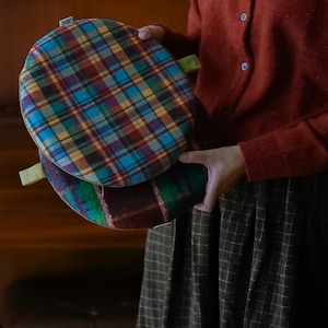 May include: Two round plaid cushions in shades of blue, red, yellow, and green are held in front of a person. Each cushion has a green fabric loop attached. The cushions have a zipper closure. The cushions are made of a soft, woven fabric.