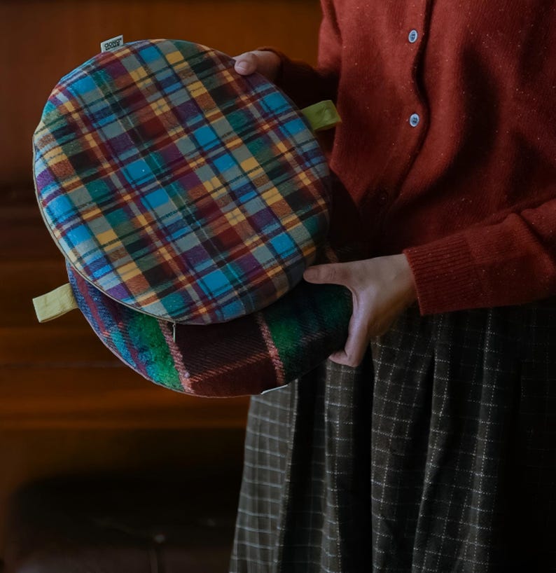 May include: Two round plaid cushions in shades of blue, red, yellow, and green are held in front of a person. Each cushion has a green fabric loop attached. The cushions have a zipper closure. The cushions are made of a soft, woven fabric.