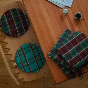 May include: Overhead shot of three round plaid cushions in green, burgundy, and pink. One cushion is on a wooden chair, two are stacked on a wooden table. A cup of coffee and a small clock are also on the table.