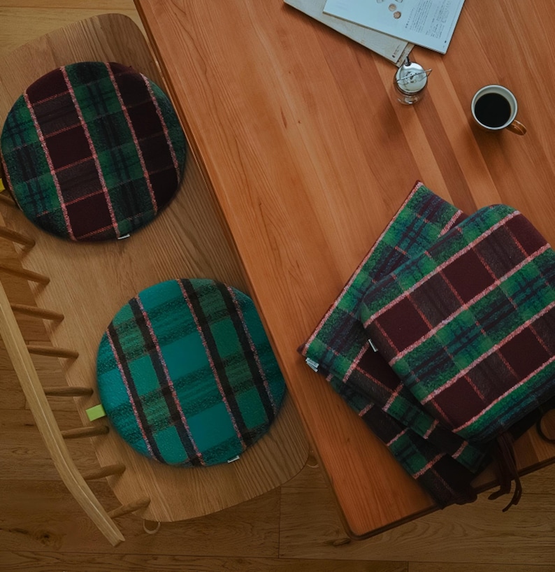May include: Overhead shot of three round plaid cushions in green, burgundy, and pink. One cushion is on a wooden chair, two are stacked on a wooden table. A cup of coffee and a small clock are also on the table.
