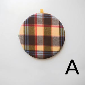 May include: A round, plaid fabric item in shades of brown, red, yellow, and blue. It has a small loop at the top and a small green tag on the side. The letter "A" is in black at the bottom right.