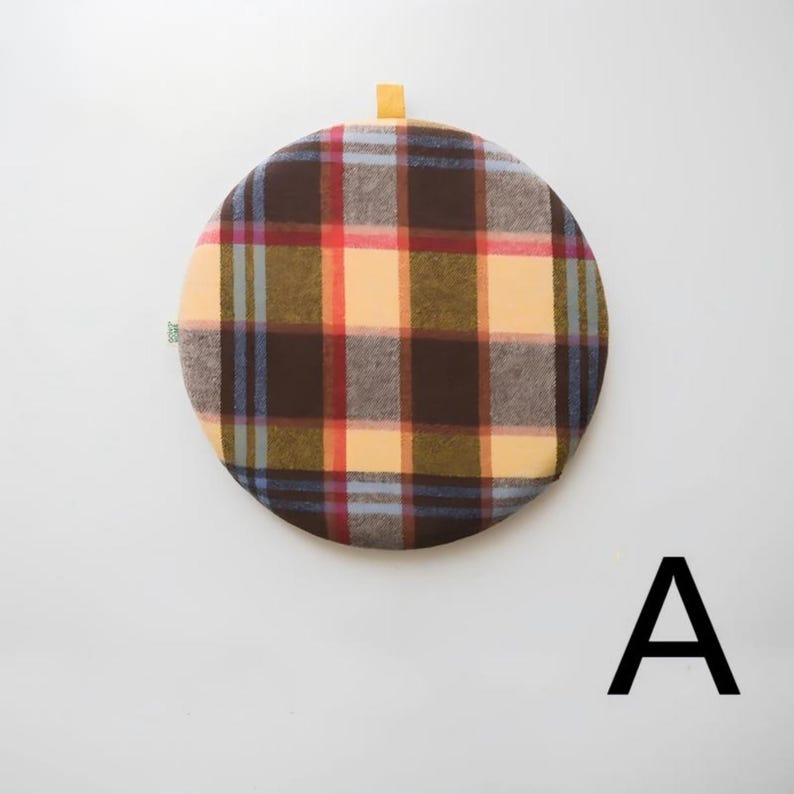 May include: A round, plaid fabric item in shades of brown, red, yellow, and blue. It has a small loop at the top and a small green tag on the side. The letter "A" is in black at the bottom right.