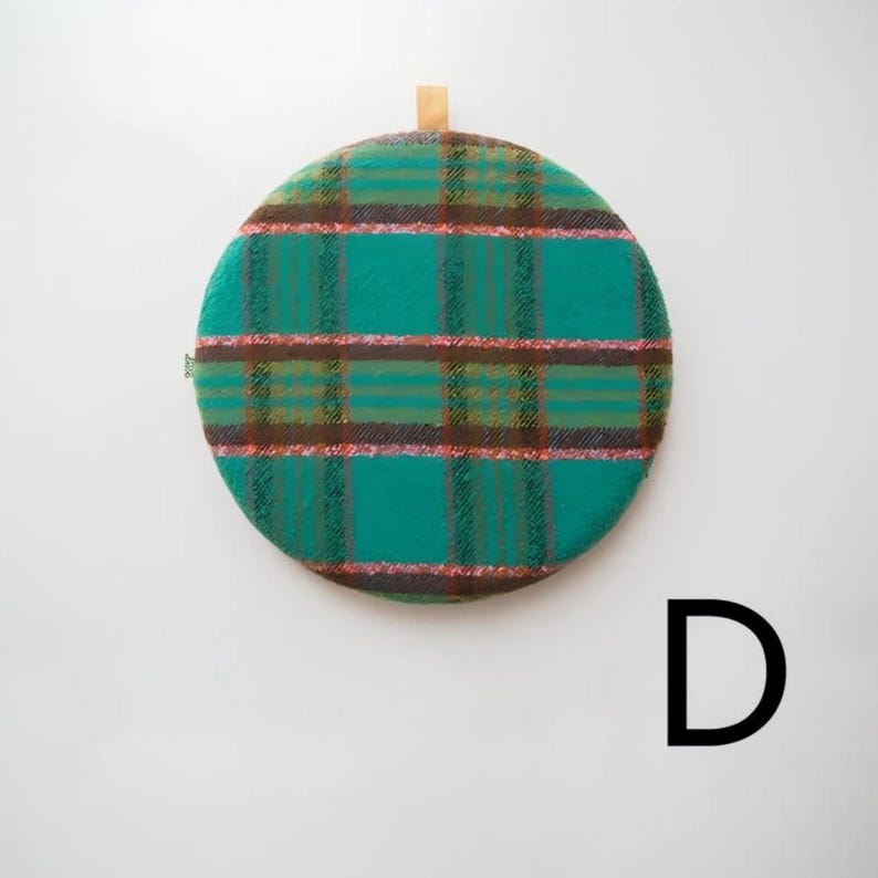May include: A round, teal and brown plaid cushion with a wooden loop for hanging. The cushion has a soft, textured appearance and a classic plaid pattern. The cushion is a decorative item.