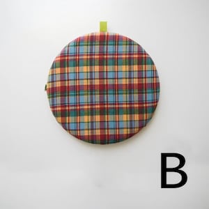 May include: A round, plaid cushion in shades of red, blue, yellow, and green. The cushion has a green loop at the top and a small green tag on the side. The cushion is set against a white background.