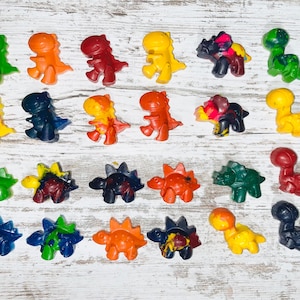 Puede incluir: Colección de ceras con forma de dinosaurio en tonos verdes, naranjas, rojos, amarillos y azules. Las ceras están dispuestas sobre una superficie de madera blanca, mostrando sus colores vibrantes y formas únicas.