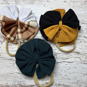 Peut inclure: Trois bandeaux pour bébés avec de grands nœuds en tissu. Un nœud est blanc avec un motif à carreaux marron, un autre est noir et or, et le troisième est vert foncé. Chaque nœud est fixé à un élastique de couleur neutre.