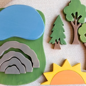 Könnte beinhalten: Ein Holzspielzeug-Set bestehend aus einer grünen Basis mit einem blauen Teich und ineinandergreifenden grauen Bergteilen. Enthalten sind zwei grüne Bäume mit braunen Stämmen, eine grüne Tanne mit braunem Stamm und eine gelb-orange Sonne mit halbrundem Kern.