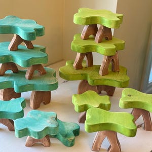 May include: Wooden toy trees in shades of green and teal. The tree tops are leaf-shaped and rest on brown wooden supports. The trees are various sizes and arranged on a white surface.