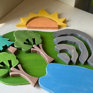May include: Wooden landscape puzzle with a green base, featuring a blue lake, grey mountains, and trees in shades of green and brown. A yellow sun with an orange centre completes the scene.