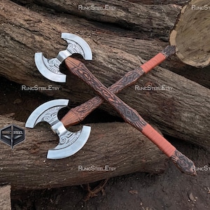 Könnte beinhalten: Zwei gekreuzte Äxte mit silbernen Klingen und aufwendig geschnitzten Holzgriffen. Die Griffe sind mit rotbraunem Leder umwickelt. Die Äxte liegen auf einem Bett aus braunen Baumstämmen. Der Etsy-Shop-Name "RunicSteelEtsy" ist sichtbar.