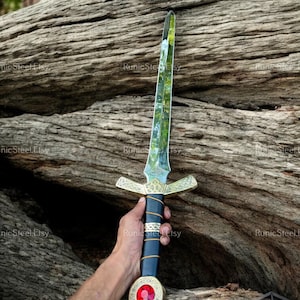May include: A decorative sword with a clear blade and a gold-colored crossguard. The handle is black with gold accents and a red jewel at the base. The sword is held against a textured wooden background.