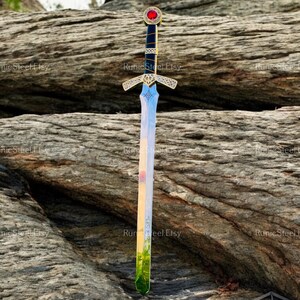 May include: A decorative sword with a gold hilt and a blade that reflects a landscape of colors. The hilt features a red gem and intricate gold detailing. The sword is displayed against a textured rock background.