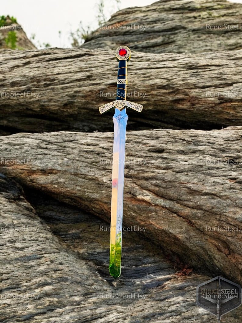 May include: A decorative sword with a gold hilt and a blade that reflects a landscape of colors. The hilt features a red gem and intricate gold detailing. The sword is displayed against a textured rock background.