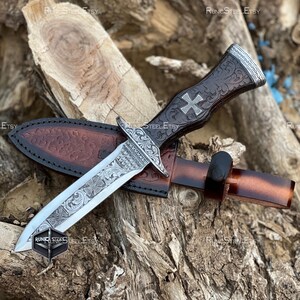 Peut inclure: Un poignard d&eacute;coratif avec une lame argent&eacute;e et un manche en cuir marron orn&eacute; d'une croix. Le poignard est dans un &eacute;tui en cuir marron. La lame et le manche pr&eacute;sentent des motifs grav&eacute;s complexes. Le poignard repose sur une surface en bois.