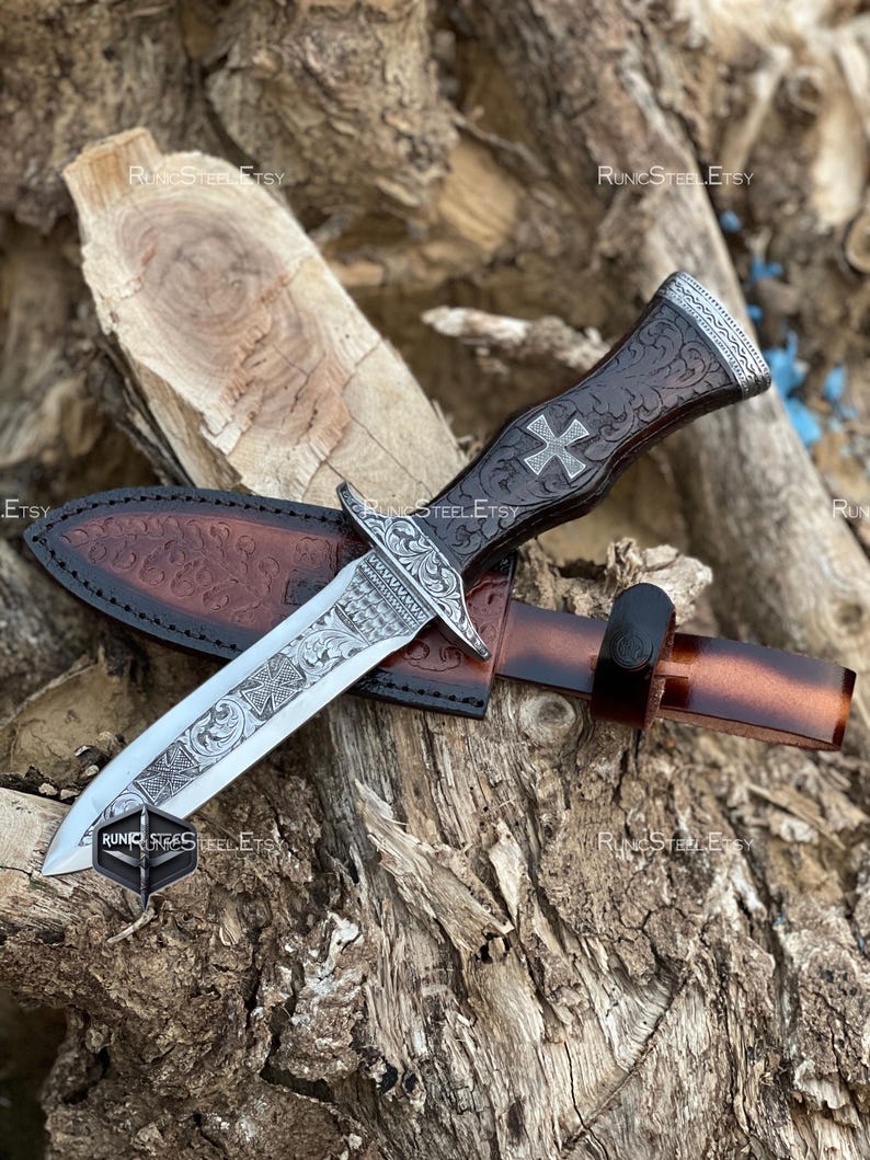 Peut inclure: Un poignard d&eacute;coratif avec une lame argent&eacute;e et un manche en cuir marron orn&eacute; d'une croix. Le poignard est dans un &eacute;tui en cuir marron. La lame et le manche pr&eacute;sentent des motifs grav&eacute;s complexes. Le poignard repose sur une surface en bois.