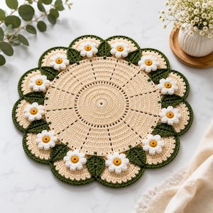 Pode incluir: Um centro de mesa redondo em crochê na cor creme com um padrão circular central e linhas radiantes. A borda é decorada com folhas de crochê verde-escuras alternadas com flores brancas tipo margarida com centros amarelos.