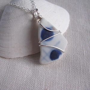 May include: A pendant necklace featuring a piece of white sea glass with two dark blue circular spots and faint blue grid lines, wrapped in silver-colored wire and suspended from a delicate silver-colored metal chain.