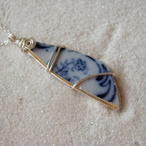 May include: A silver necklace featuring a pendant made from a piece of blue and white porcelain. The porcelain has a floral design and is wrapped in silver wire. The necklace is displayed on a sandy surface.