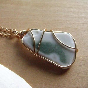 May include: A pendant necklace featuring a smooth, white, and green sea glass piece wrapped in gold-toned wire. The pendant is attached to a delicate gold-toned metal chain with small circular links, creating a simple and elegant jewelry piece.