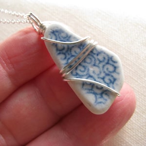 May include: A pendant necklace featuring a piece of white sea pottery with a blue swirling pattern, secured in a silver-colored wire wrap. The pendant hangs from a thin silver-colored chain, showcasing the smooth, weathered texture of the ceramic.