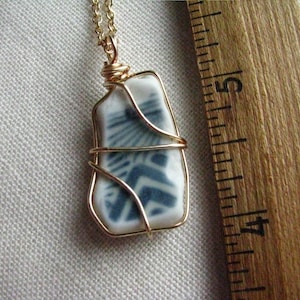 May include: A pendant featuring a white ceramic shard with a blue geometric pattern, encased in gold-colored wire wrapping and attached to a gold-colored chain, positioned next to a wooden ruler showing a length of approximately 1.25 inches.